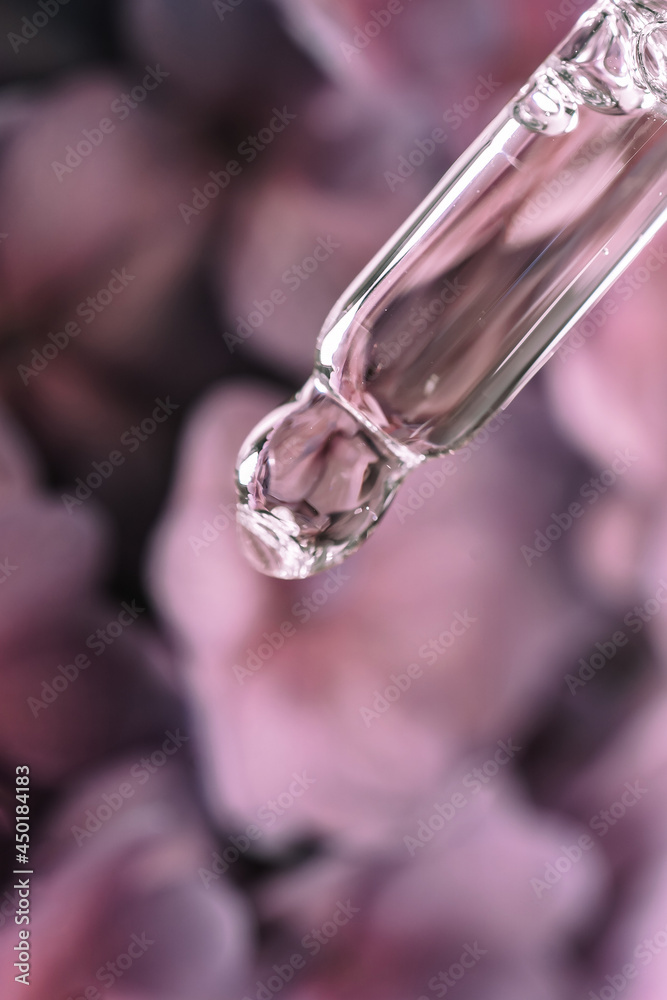 A drop of cosmetic oil falls from the pipette on a purple floral background. A drop of cosmetic oil falls from the pipette on a purple floral background.