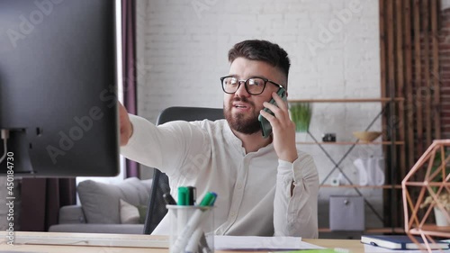 Portrait shot Handsome young man office worker in glasses speaking on the mobile phone and explaining something of working process while looking at the computer screen. Freelancer work in home office.