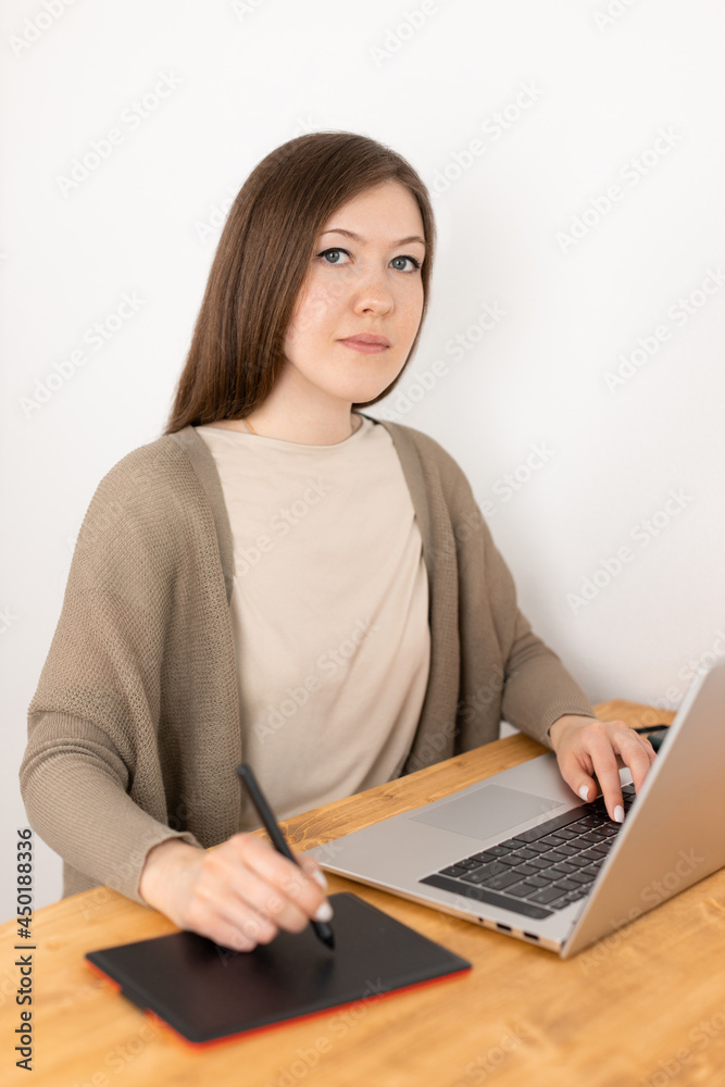 Pretty caucasian woman graphic designer sitting at workspace and looking into camera. Girl in beige t-shirt and grey cardigan work from home office. On the wooden desk laptop and digital tablet.