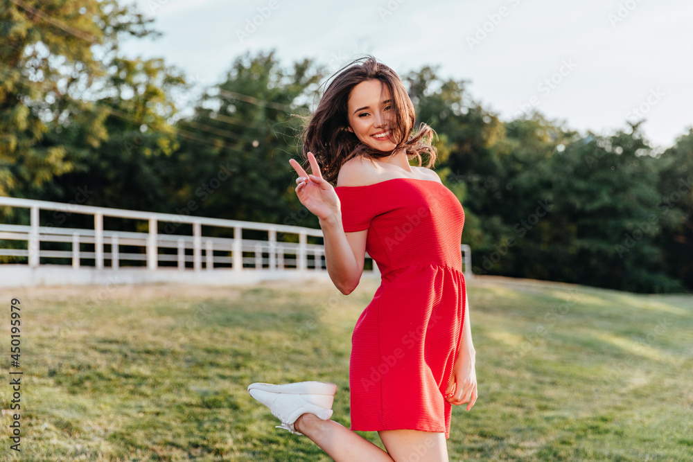 Shapely lovable girl in red outfit posing in park with pleasure. Spectacular brunette woman in dress standing on nature background with peace sign. Shapely lovable girl in red outfit posing in park with pleasure. Spectacular brunette woman in dress standing on nature background with peace sign.