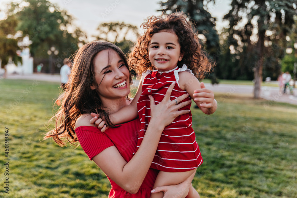 Excited female model holding little girl and smiling on nature background. Outdoor photo of spectacular white woman fooling around with daughter during rest in park.