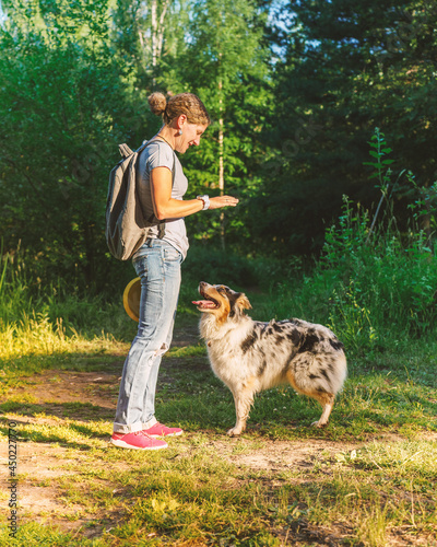 Full length of smiling happy woman in casual clothes playing with lovely dog in park on summer day, female pet owner giving command to stay to young Australian Shepherd during obedience training 