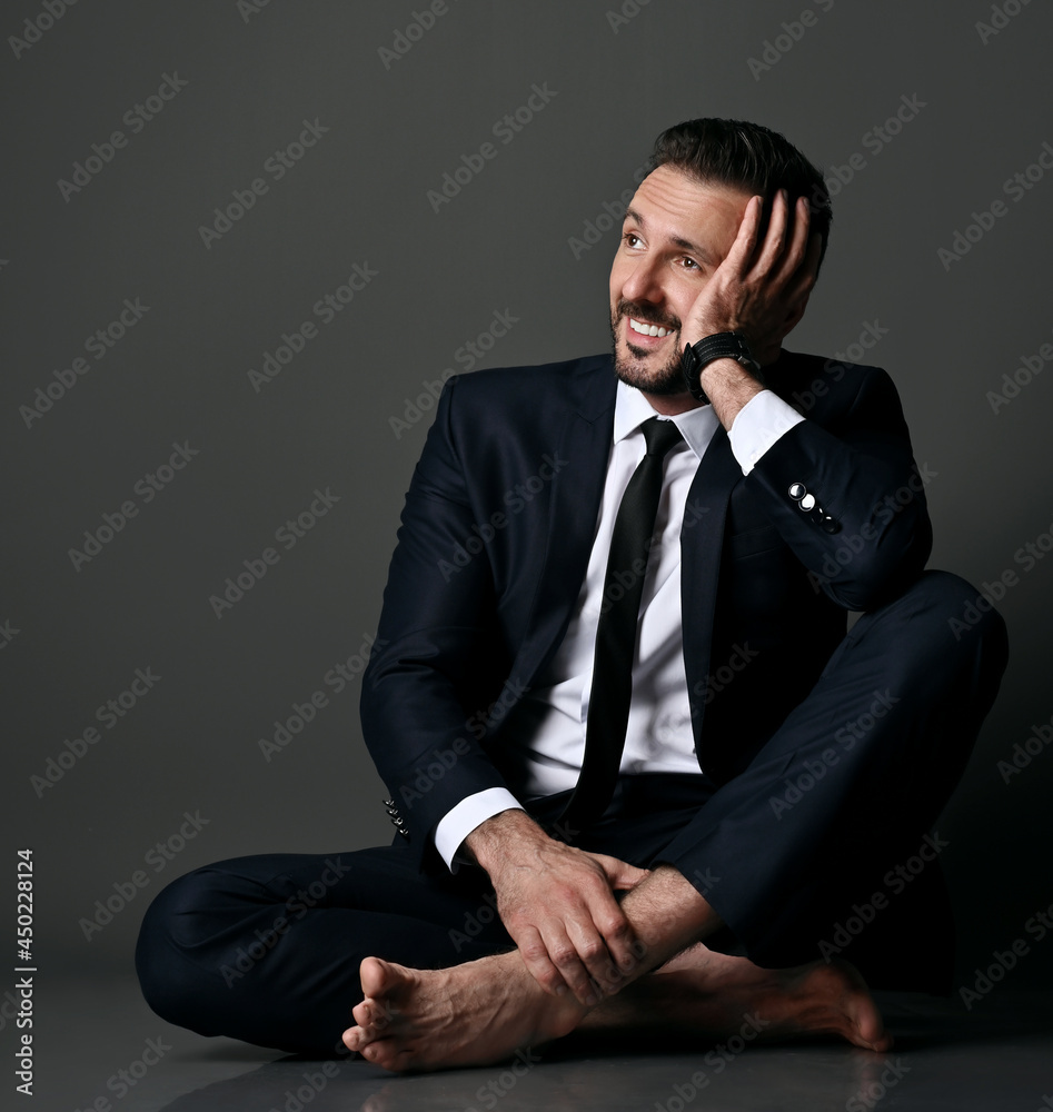 Silly smiling adult bearded man, failed manager, broker in black office suit has just seen his mistake, sits barefooted on floor with legs crossed holding hand at head, looking aside at copy space Silly smiling adult bearded man, failed manager, broker in black office suit has just seen his mistake, sits barefooted on floor with legs crossed holding hand at head, looking aside at copy space