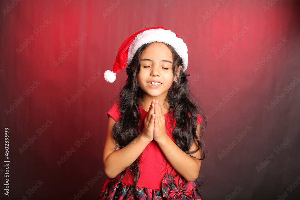 a beautiful little dark-haired girl in a red dress and a Santa Claus hat is surprised and makes a wish for the new year a beautiful little dark-haired girl in a red dress and a Santa Claus hat is surprised and makes a wish for the new year