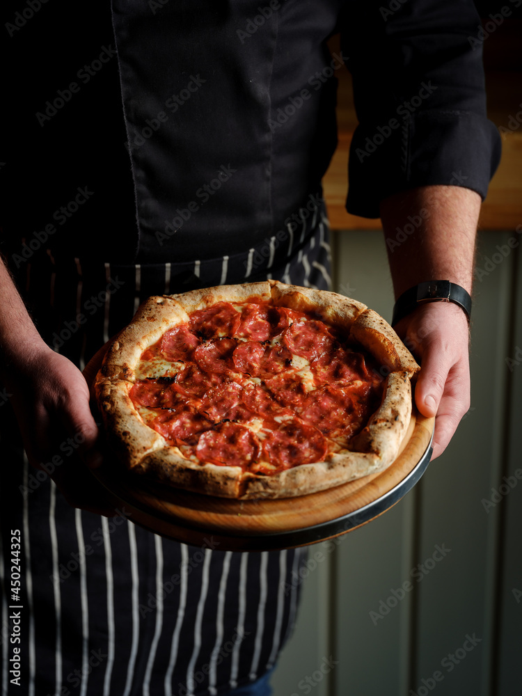 Italian traditional pizza bread with pepperoni meat and red tomatoes sauce in cook baker hands on dark restaurant background 