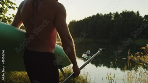 Woman swimming, sitting on surfing board in sunlight, while man surfer going to river with surfboard and paddle. Couple preparing to surf at sunrise. Male coach goes down to water holding sup board.