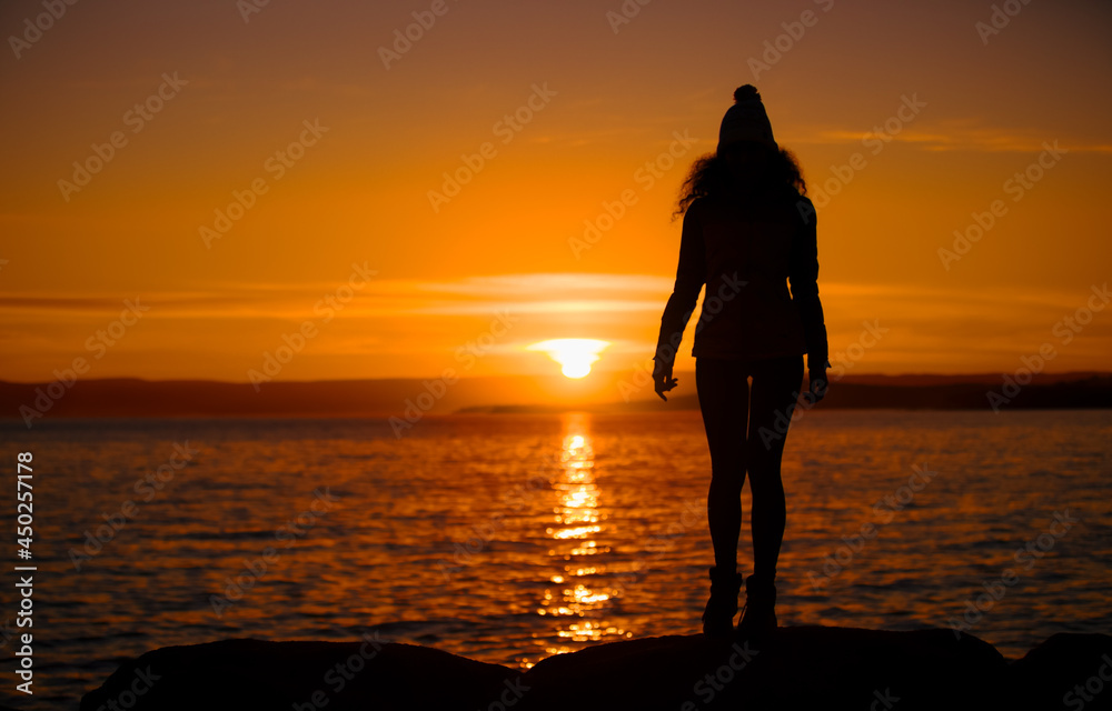 Silhouette of a young lady enjoying freedom by the sea at sunset. Slim relaxing woman in the evening looking away. Portrait Photography