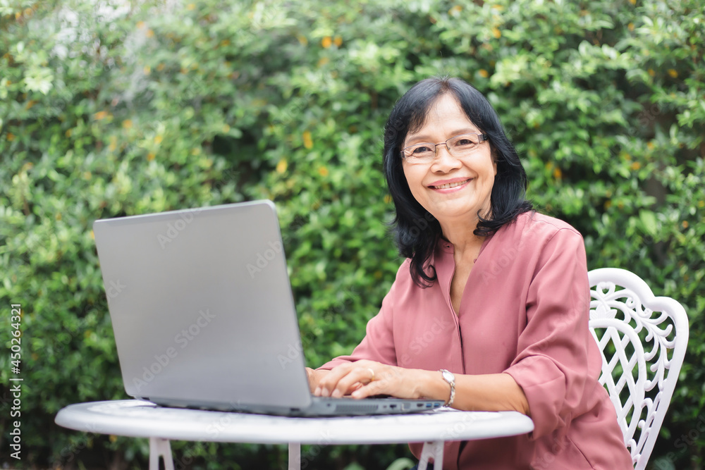 Senior Asian woman playing laptop in garden at home. Mature middle aged female in glasses working online with computer and looking at camera with green background. Senior Asian woman playing laptop in garden at home. Mature middle aged female in glasses working online with computer and looking at camera with green background.