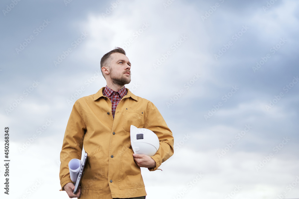 Supervising on site concept. Serious cute caucasian bearded construction contractor, supervisor, foreman or architect with blueprints, laptop and helmet in hands at construction site in orange jacket
