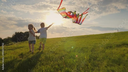 Happy smiling little children running holding hands and playing with a kite on the meadow in the rays of the sun at sunset
