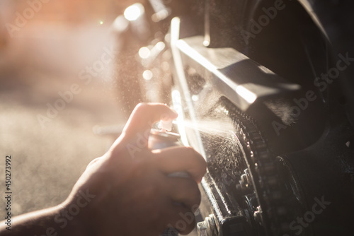 Outdoor sun reflect photograph of hand applying spray oil on motorbike chain for maintenance.