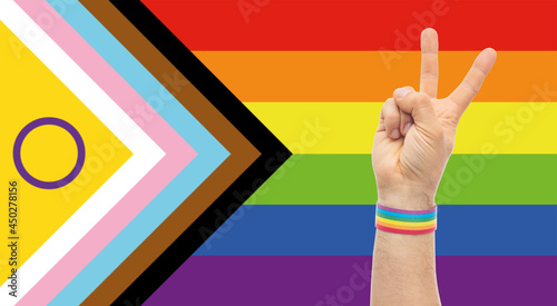 lgbtq, trans and intersex rightsl concept - close up of male hand wearing rainbow wristband showing peace sign over progress pride flag on background