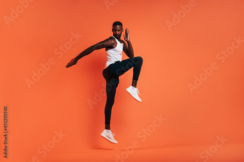 Muscular athlete jumping in the air. Man in sports clothes doing fitness workout over orange background.