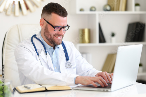Positive professional male physician therapist in white coat with stethoscope working on a laptop