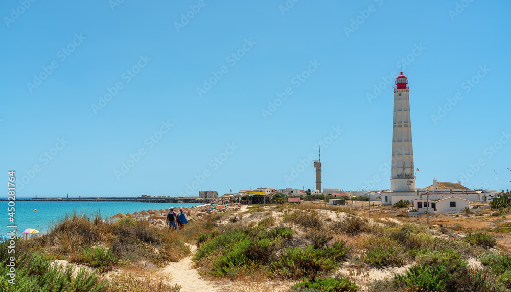 View of the beach of the Portuguese island of Farol with a lighthouse and a couple walking along the path. Ria Formosa