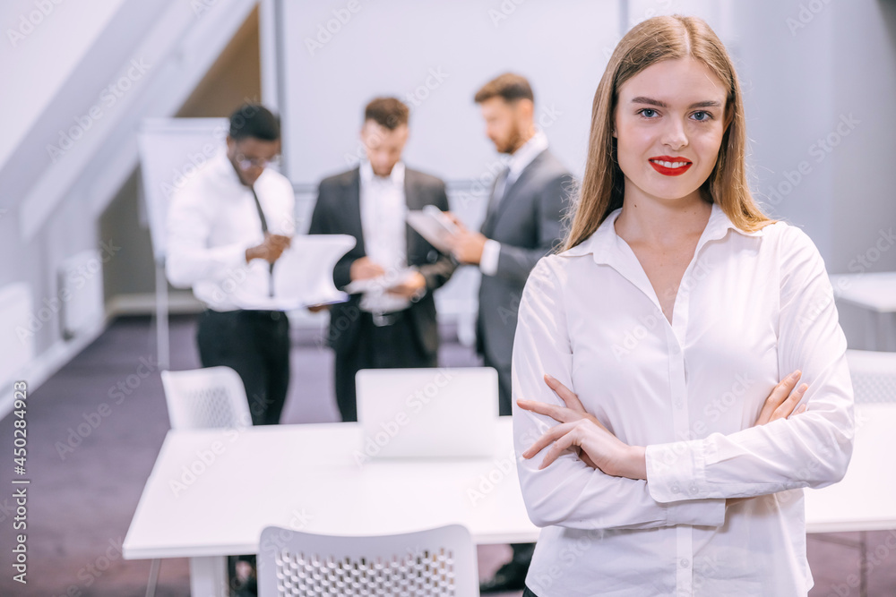Beautiful young smiling professional woman in office with folded hands and confident expression as other workers have a meeting in the background Beautiful young smiling professional woman in office with folded hands and confident expression as other workers have a meeting in the background