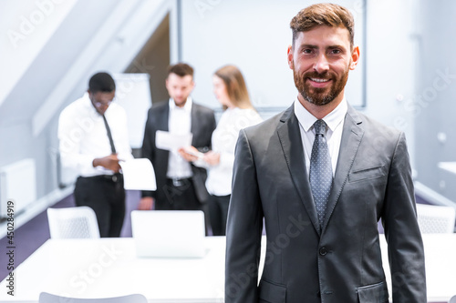 Happy businessman standing in office with colleagues in the background working at table
