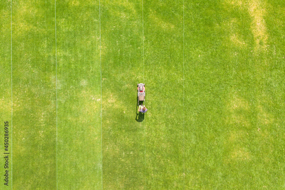 Obraz premium Aerial view of small figure of man worker trimming green grass with mowing mashine on football stadium field in summer.