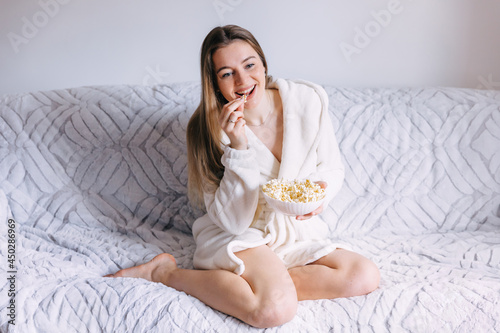 Young woman in white bathrobe with popcorn watching movie on sof