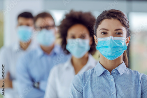 Multicultural group of business people with face masks on standing in office with arms crossed and looking at camera. Selective focus on woman in the foreground.