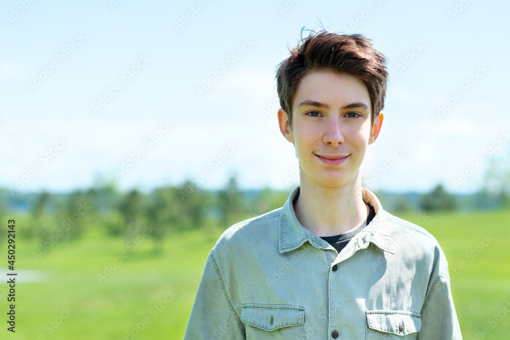 Handsome cheerful teen boy 15 years old smiling and looking at camera against blue sky and green grass at spring meadow.