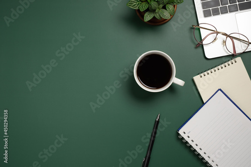 Desk office with laptop, blank notebook, flower, coffee cup and pen on green table. Flat lay top view copy space. Home office.