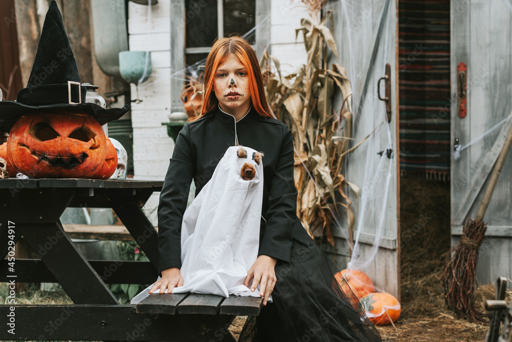 girl in a witch costume with a dog in a ghost costume having fun on the porch of a house decorated to celebrate a Halloween party