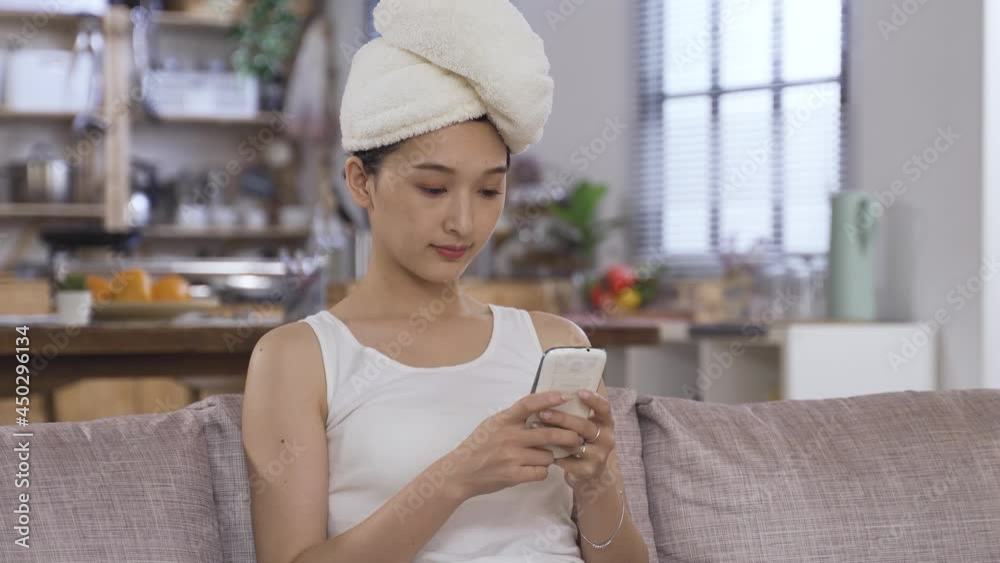 portrait asian lady wrapping her hair with a towel is sliding on the phone screen and sending messages at leisure on living room couch. portrait asian lady wrapping her hair with a towel is sliding on the phone screen and sending messages at leisure on living room couch.