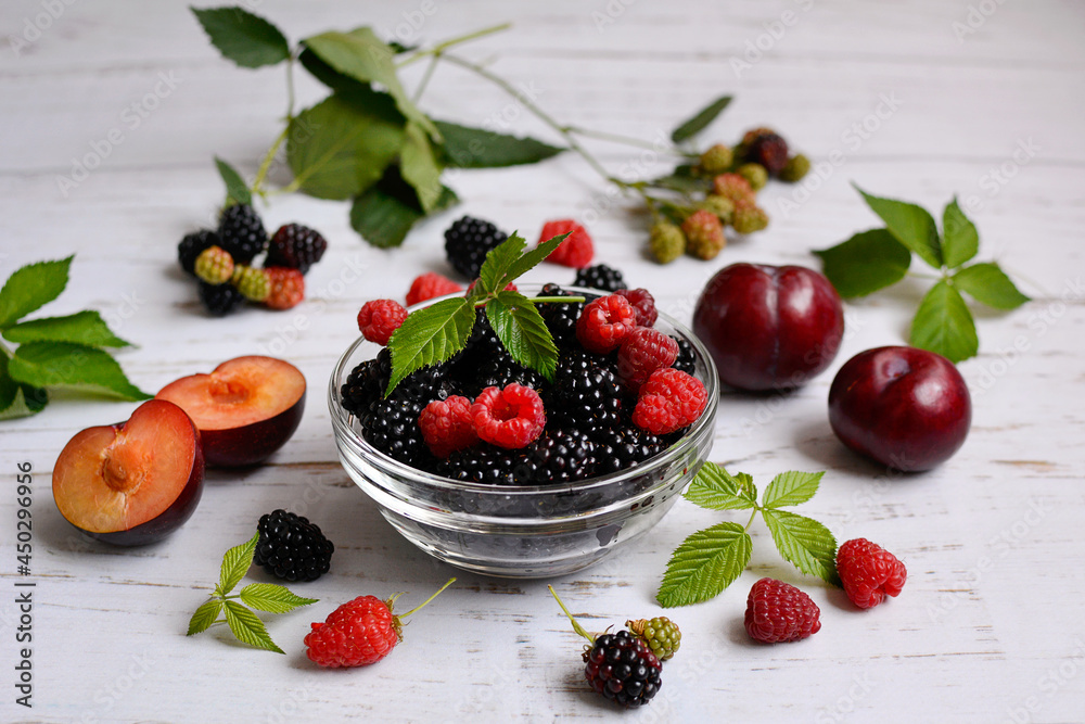 The concept of a juicy and vitamin summer. Ripe raspberries, blackberries, plums and mint leaves on a light background, close-up The concept of a juicy and vitamin summer. Ripe raspberries, blackberries, plums and mint leaves on a light background, close-up