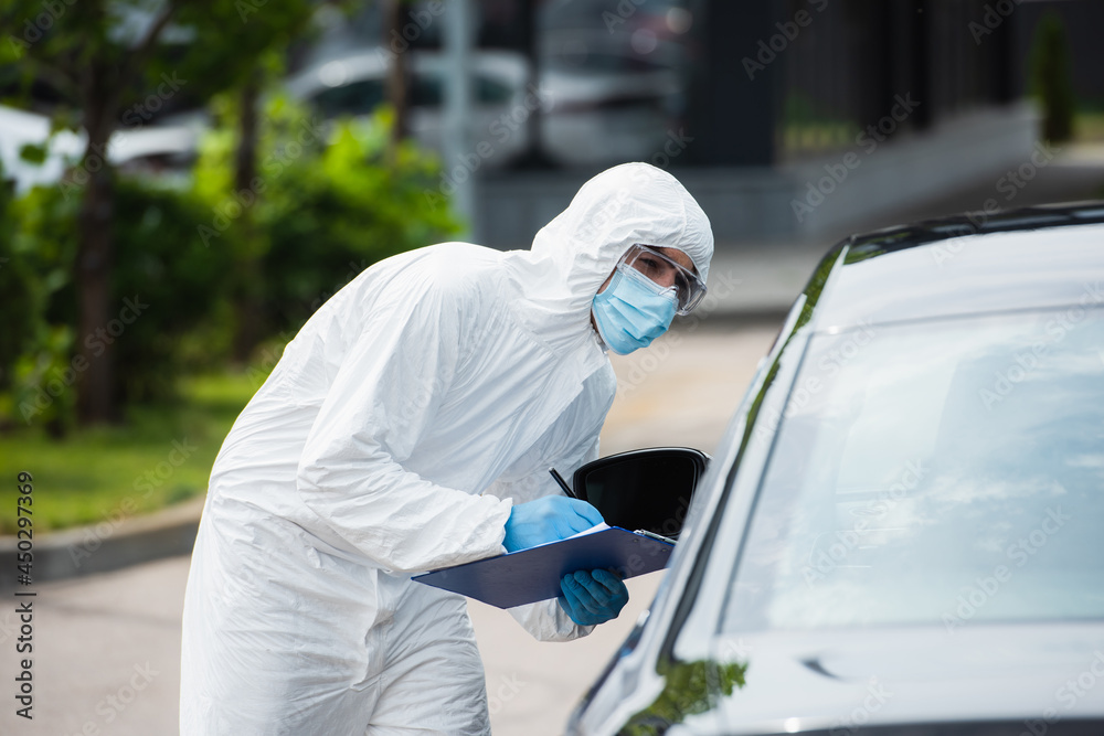 Border guard in medical mask with clipboard looking through car on blurred foreground