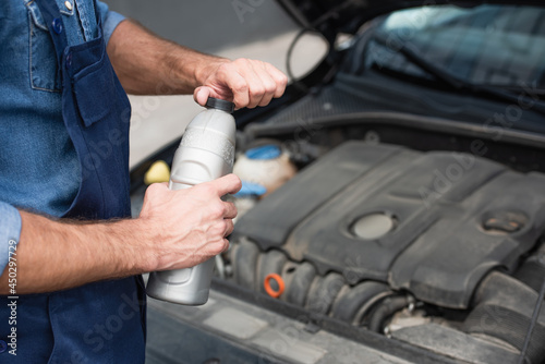 Cropped view of mechanic in uniform holding bottle of motor oil near blurred car