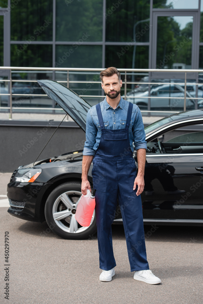 Repairman in overalls holding windshield washer fluid near auto outdoors
