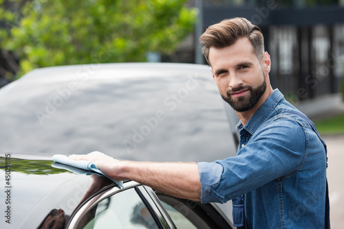 Mechanic looking at camera while waxing car with rag outdoors