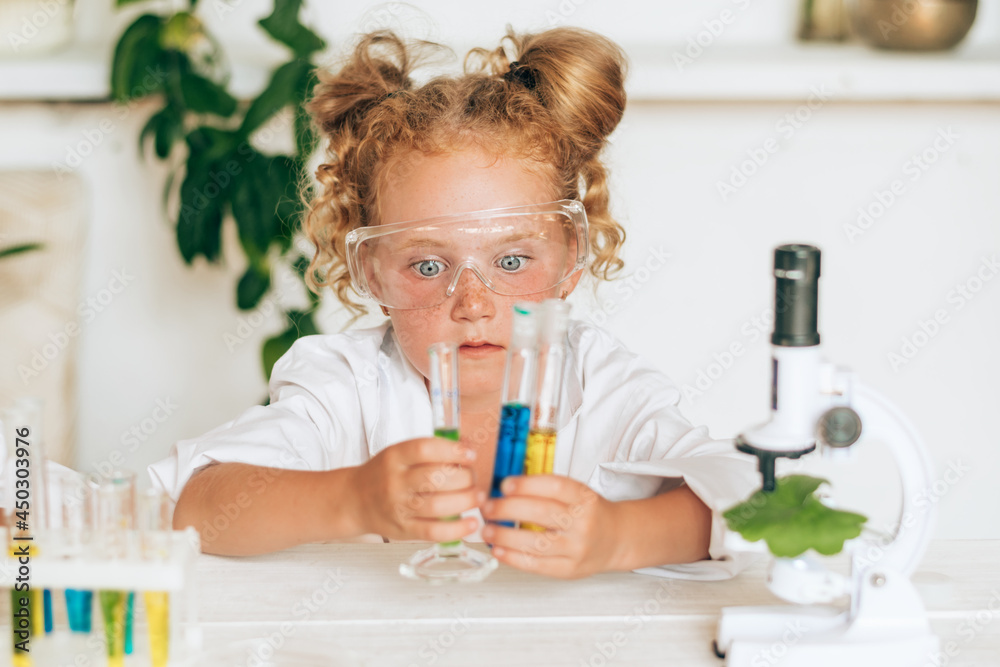 Surprised little red-haired girl in white uniform conducting chemical experiments in a laboratory.Back to school concept.Young scientists.Natural sciences.Preschool and school education of children. Surprised little red-haired girl in white uniform conducting chemical experiments in a laboratory.Back to school concept.Young scientists.Natural sciences.Preschool and school education of children.