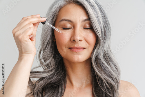 Mature shirtless woman with grey hair applying face serum