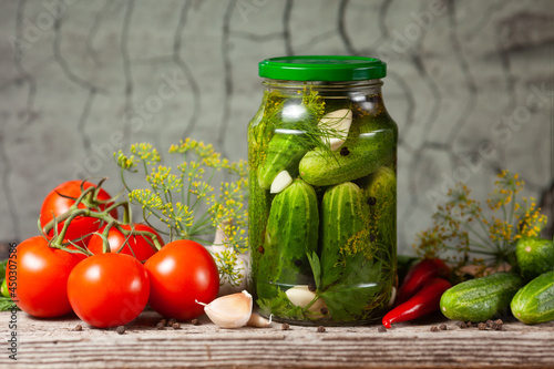 Pickled cucumbers in glass jars and spices and vegetables for preparation of pickles on wooden background