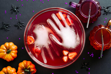 Halloween party concept. Bowl with bloody drink, frozen hands, strawberries and halloween decorations on dark background. Flat lay, top view.