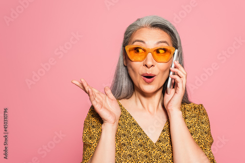 surprised middle aged woman in yellow sunglasses talking on cellphone and pointing with hand isolated on pink