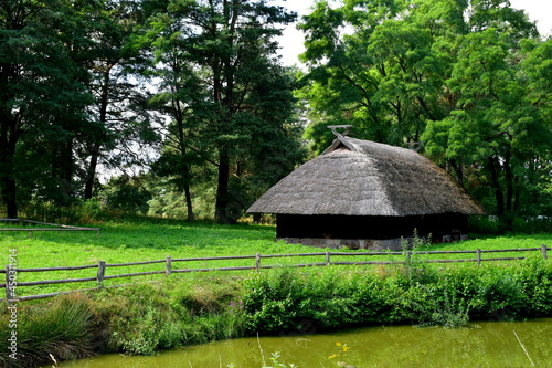 A close up on a wooden shack, hut, or house standing next to a small pond, lake, or swamp covered with shrubs, reeds and other flora seen on a sunny summer day near a dense forest or moor in Poland