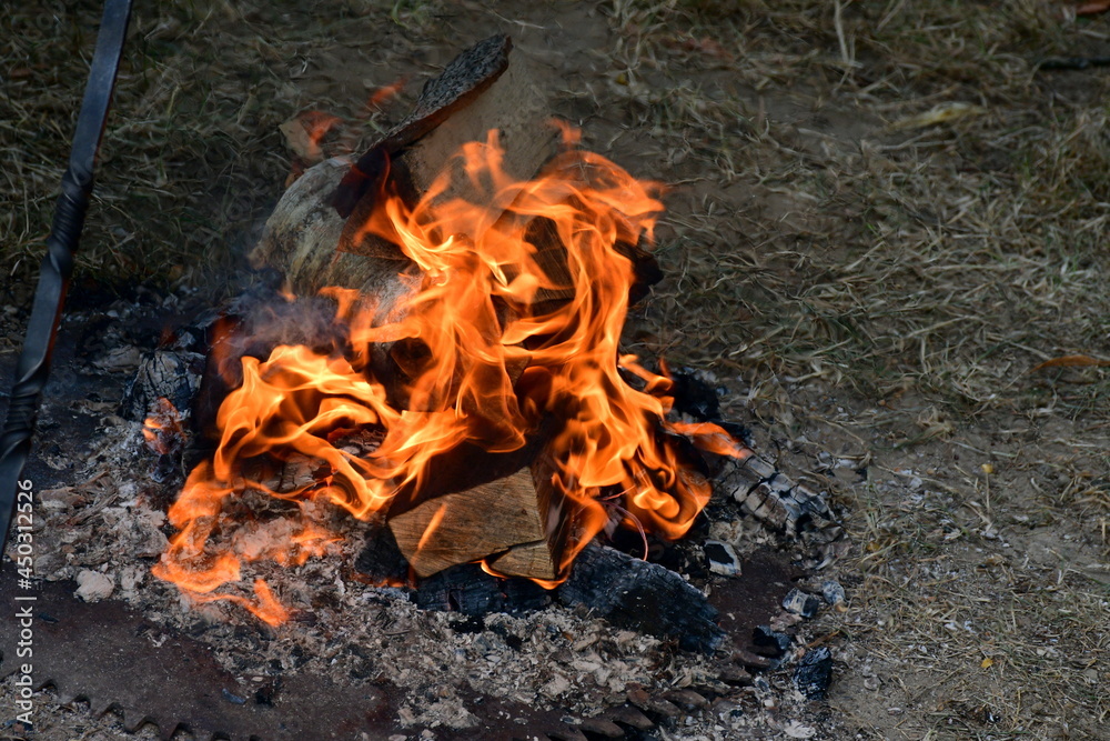 A close up on a bonfire with hot flames coming from flaming logs arrenged around a metal element with some ash below the fire and some sandy surface nearby seen on a sunny summer day in Poland A close up on a bonfire with hot flames coming from flaming logs arrenged around a metal element with some ash below the fire and some sandy surface nearby seen on a sunny summer day in Poland
