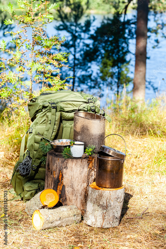 Backpack of traveller, bowler pot, enameled mug with coffee or tea, on wooden stump in the forest. Lunch break for rest during hiking or tracking travel. Concept of active tourism in summer in Karelia Backpack of traveller, bowler pot, enameled mug with coffee or tea, on wooden stump in the forest. Lunch break for rest during hiking or tracking travel. Concept of active tourism in summer in Karelia