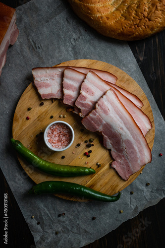 Slices of fatty bacon on a round wooden cutting board  with two green peppers,  pink salt, peppercorns and some bread on a piece of parchment paper