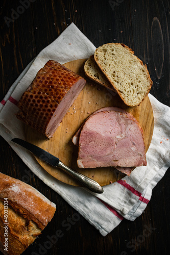 Slices of ham on a round wooden cutting board  with silver knife and some white bread on white kitchen towel and wooden table