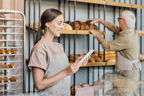 Happy female clerk looking through prices for bread against mature man putting loaves on display