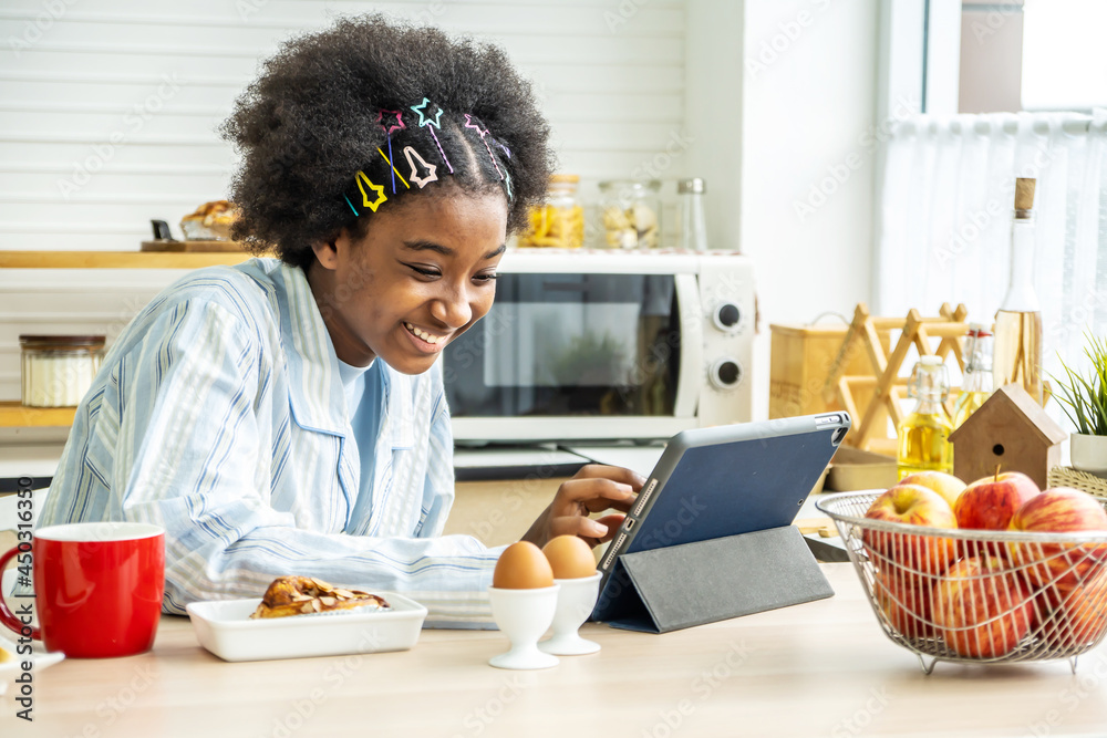 Obraz premium Young woman african american in the kitchen while having breakfast and sipping tea or milk in the cup send a message or calls with the tablet and smiles, Concept of social network, message, technology