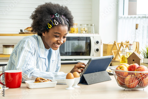 Young woman african american in the kitchen while having breakfast and sipping tea or milk in the cup send a message or calls with the tablet and smiles, Concept of social network, message, technology