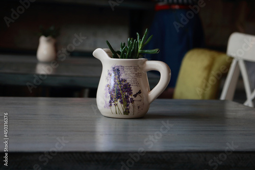 Cute milk jug with some green plants and a painting of lavender on a gray table. 