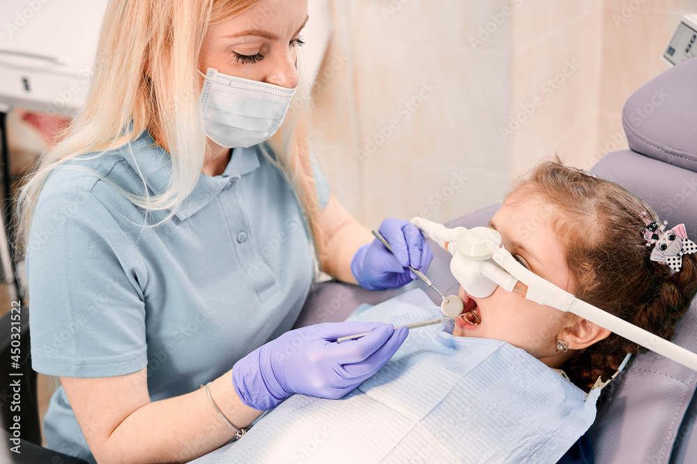 Female dentist checking child teeth with dental explorer and mirror while girl lying in dental chair with inhalation sedation at dental office. Concept of pediatric, sedation dentistry and dental care
