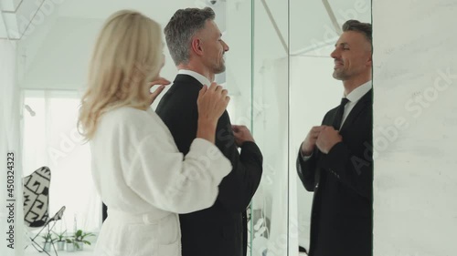 Mature woman adjusting suit of her husband while standing in front of the mirror