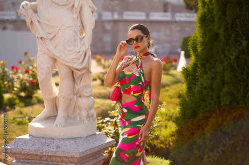 Young elegant fashionable woman wearing colorful dress, sunglasses and holding pink handbag posing in beautiful garden on a sunny summer day photo